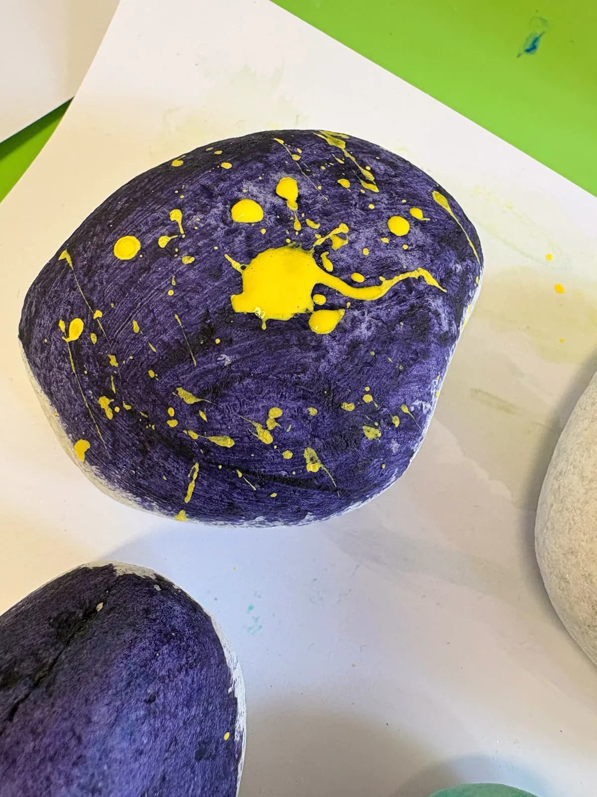 A pebble painted deep purple and splattered with yellow paint, drying on paper.